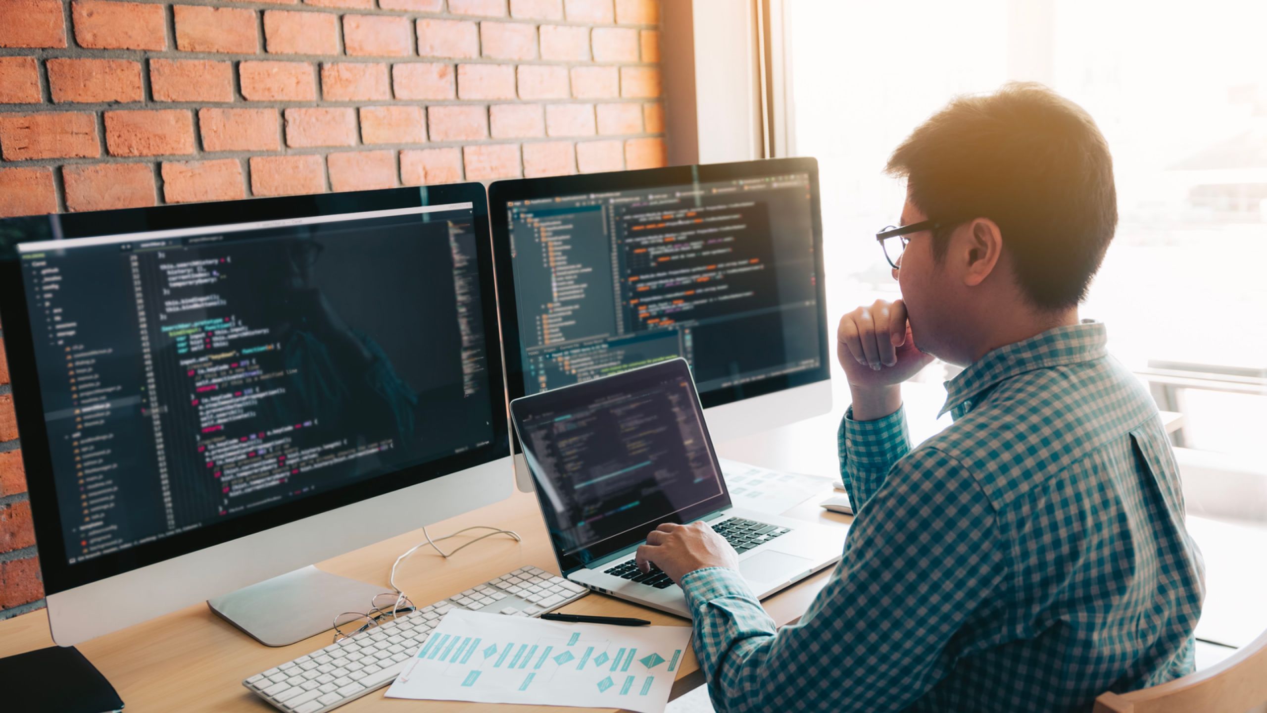 A man with glasses working on a laptop with two large computer monitors behind it